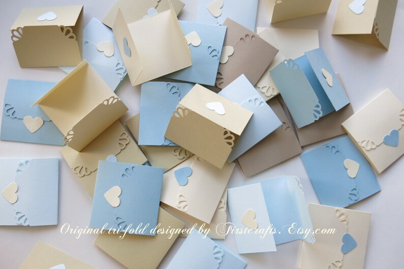 May include: A collection of folded cards with a heart cut-out design. The cards are in shades of blue and beige. The text "Original trifold designed by Trisicrafts. Etsy.com" is visible at the bottom of the image.