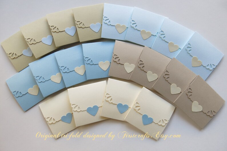 May include: A set of 18 tri-fold favor boxes in three colors: cream, light blue, and beige. Each box features a cut-out design of a heart with a scalloped edge. The boxes are perfect for holding small gifts or favors.