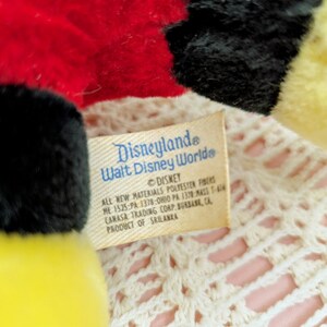 Puede incluir: Un peluche amarillo, negro y rojo con una etiqueta blanca que dice "Disneyland Walt Disney World&reg; DISNEY ALL NEW MATERIALS POLYESTER FIBERS ME 1525-PA 1378-OHIO PA 1378-MASS 1-416 CANASA TRADING CORP. BURBANK, CA PRODUCT OF SRI LANKA"