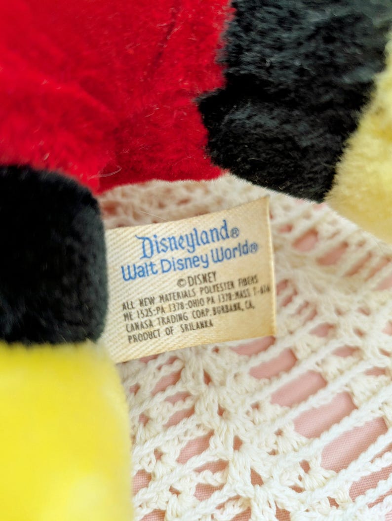 Puede incluir: Un peluche amarillo, negro y rojo con una etiqueta blanca que dice "Disneyland Walt Disney World&reg; DISNEY ALL NEW MATERIALS POLYESTER FIBERS ME 1525-PA 1378-OHIO PA 1378-MASS 1-416 CANASA TRADING CORP. BURBANK, CA PRODUCT OF SRI LANKA"