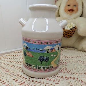 Peut inclure: Pichet à lait en céramique blanche avec deux petites poignées. Le pichet présente une scène pastorale avec une bordure florale rose. La scène représente un champ vert, des arbres, un lac et une route sinueuse. Un objet décoratif.