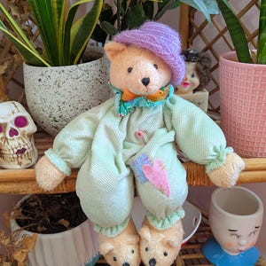 Könnte beinhalten: Plüsch-Teddybär-Puppe mit lila Strickmütze und grünem Outfit, mit bärenförmigen Hausschuhen. Die Puppe sitzt auf einem Bambusregal, umgeben von Topfpflanzen und dekorativen Gegenständen.