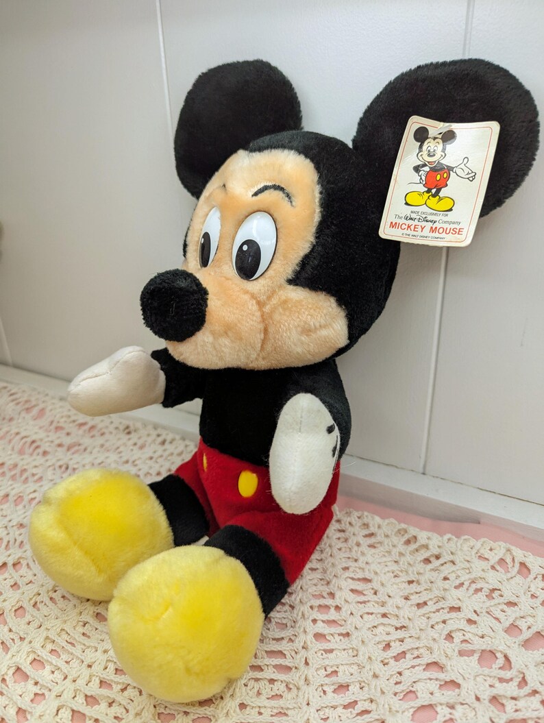 Puede incluir: Un juguete de peluche de Mickey Mouse con orejas negras, nariz negra y ojos grandes. Lleva pantalones cortos rojos con un bot&oacute;n amarillo y zapatos negros. El juguete tiene una etiqueta que dice "The Walt Disney Company Mickey Mouse".