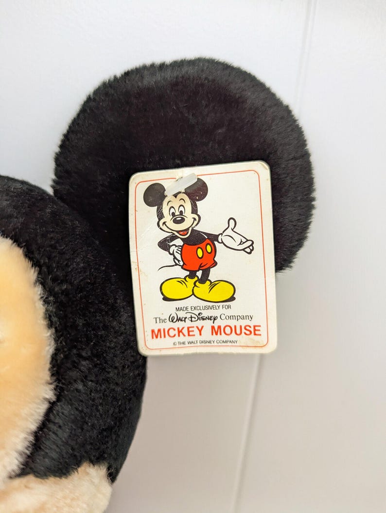 Puede incluir: Un juguete de peluche de Mickey Mouse con un dise&ntilde;o en blanco y negro. El juguete tiene una etiqueta que dice "Hecho exclusivamente para The Walt Disney Company Mickey Mouse &copy; The Walt Disney Company".