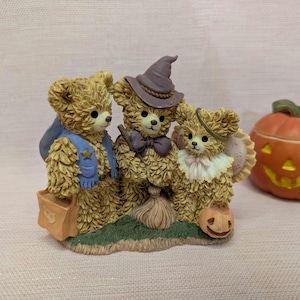 May include: A Halloween-themed figurine of three teddy bears in costume. One bear is dressed as a witch with a hat and broom, another wears a cape and carries a bag, and the third has a halo and a pumpkin. A carved pumpkin is also visible.