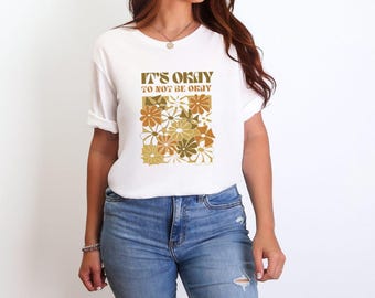Puede incluir: Camiseta blanca con un diseño floral marrón y amarillo y el texto "It's okay to not be okay."