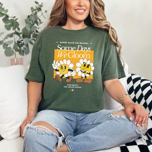 May include: Green t-shirt with a graphic of two cartoon flowers with the text "Some Days We Gloom" and "For Both We Leave Room".