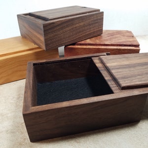 May include: Three wooden boxes with lids, one open, showing a black velvet interior. The boxes are made of different types of wood, including walnut and cherry.