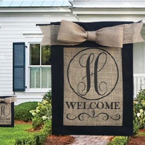 May include: A burlap welcome sign with a black border and a large letter "H" in a circle. The sign has a brown bow and the words "Welcome" underneath the circle.