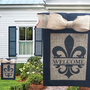 May include: Two burlap welcome flags with a black background and a black fleur-de-lis design. The larger flag has a burlap bow and the smaller flag is on a black metal stand. Both flags say "Welcome" in black lettering.
