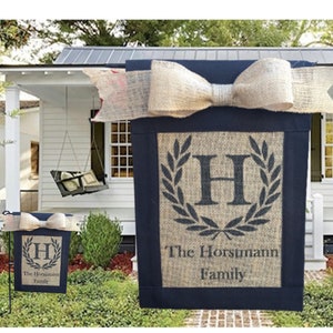 May include: A navy blue and burlap family garden flag with a large bow at the top. The flag features a monogram letter "H" within a laurel wreath and the family name "The Horstmann Family". A smaller version is also displayed.