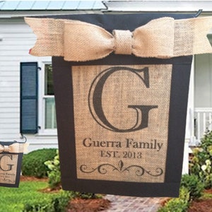 May include: A burlap garden flag with a black border and a large letter "G" in the center. The flag also has the text "Guerra Family EST. 2013" and a decorative scroll design.