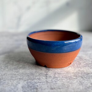 May include: A small, handmade ceramic bowl with a two-tone design. The lower portion is a natural terracotta color, while the upper rim features a glossy, deep blue glaze. The bowl has a simple, rounded shape and is suitable for various uses.