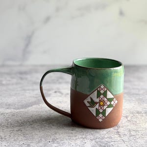May include: A handmade ceramic mug with a green glazed top and handle, and a brown base. The mug features a decorative square design with a hummingbird and geometric patterns. The mug is on a gray surface.
