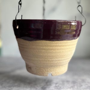 May include: A hanging ceramic planter with a two-tone design. The top is a glossy, deep purple, while the lower portion has a textured, beige finish. The planter is suspended by a black metal chain, perfect for displaying plants.