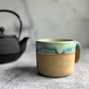 May include: A handmade ceramic mug with a tan base and a teal and cream-colored rim. The mug has a rounded shape and a small handle. A black teapot is slightly visible in the background.