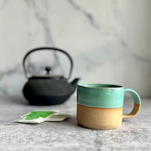 May include: A handmade ceramic mug with a teal top and tan bottom. A black teapot and tea packets are in the background. The mug has a curved handle and is likely used for tea or coffee.