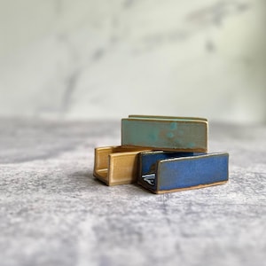 May include: A stack of four ceramic business card holders in shades of blue, gold, and teal. Each holder is rectangular with a small slot to hold cards. The holders have a glossy finish and are stacked on a gray surface.