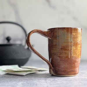 May include: A handmade ceramic mug with a brown and gold glaze. The mug has a curved handle and a rustic appearance. A black teapot and tea bags are in the background, suggesting a tea-drinking theme.