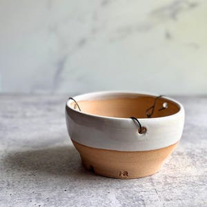May include: A round ceramic hanging planter with a glossy white glazed upper section and a matte terracotta-colored lower base. The bowl features three small holes along the rim for suspension wires and the stamped letters HR on the side.