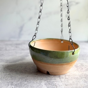 May include: A hanging ceramic planter with a two-tone design. The upper portion is glazed in a glossy green, while the lower section is a natural tan color. The planter is suspended by black metal chains, perfect for displaying plants.