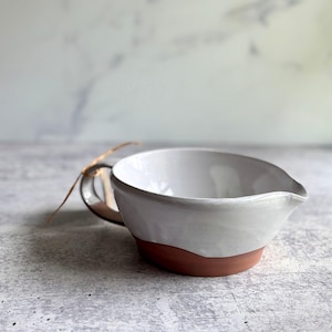 May include: A ceramic pitcher with a glossy white glazed interior and upper exterior, contrasted by a raw, unglazed terracotta-colored base. The vessel features a curved handle with a thin, light-colored string tied around it and a small spout.