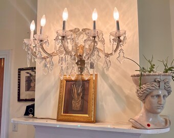 May include: A wall-mounted crystal chandelier with five candle-style lights, featuring clear glass prisms and metal arms. Below it sits a framed rectangular artwork in a gold-toned frame and a light-colored stone bust planter with green foliage.