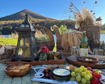 May include: A picnic scene with a variety of food and drink items. A decorative lantern, baskets, and wooden trays are arranged on a green blanket. Includes a bottle of apple cider, crackers, fruit, and pesto.