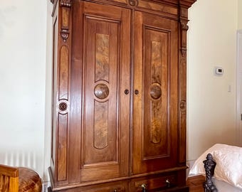 May include: A tall, dark brown wooden wardrobe featuring two large doors with vertical burl wood panels and central circular carvings. The piece includes two bottom drawers with small dark knobs, decorative crown molding, and small white caster wheels.