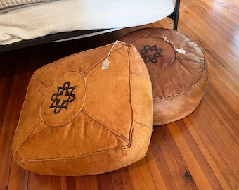 May include: Two tan leather poufs with stitched panel construction and a dark brown geometric emblem centered on the top surface. Each pouf features a small white paper tag attached with a string, showcasing a textured finish and visible seams.