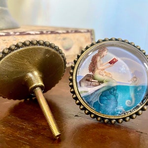 May include: Two bronze-toned cabinet knobs. One knob features a glass dome with a mermaid reading a book, a seahorse, and a blue and green underwater scene. The other knob is plain with a beaded edge.