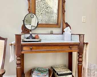 May include: A wooden washstand featuring a rectangular mirror with a distressed finish, a white marble top, and a single drawer. The piece includes a lower shelf holding a wooden bowl with magazines and a patterned box, plus side towel bars.