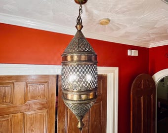 May include: A hanging pendant lamp made of brass with an intricate, perforated metal pattern. The cylindrical fixture features a pointed top and bottom, suspended from the ceiling by a chain, with light emitting through the cut-out metal design.