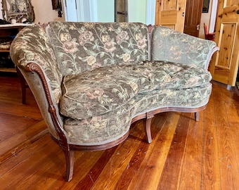 May include: A curved loveseat featuring a dark green fabric upholstery with a multi-colored floral pattern. The piece has a wooden frame with carved detailing along the arms and legs, a tufted seat cushion, and a rounded backrest design.