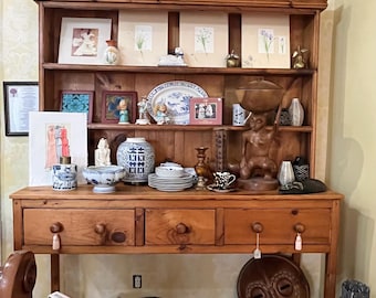 May include: A large wooden hutch with three drawers and an open shelf base, displaying a collection of ceramic bowls, plates, vases, framed art prints, a wooden sculpture, a woven basket, a carved wooden mask, and a decorative metal cross.