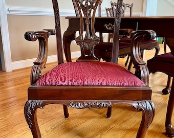 May include: A dark wood armchair featuring a carved splat back with intricate scrollwork and foliage details. The seat is upholstered in a deep red fabric with a textured pattern. The chair has curved arms and cabriole legs with claw feet.