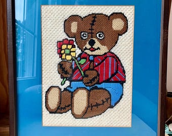 May include: Framed cross-stitch art featuring a brown teddy bear holding a colorful flower. The bear wears a red and blue striped shirt and blue pants. The artwork is framed in a dark brown frame with a blue mat.