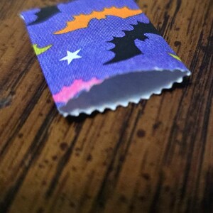 May include: A small rectangular paper pouch with a purple background featuring a repeating pattern of black bats, orange bats, white stars, and lime green accents. The opening of the pouch has a decorative scalloped edge along the top rim.