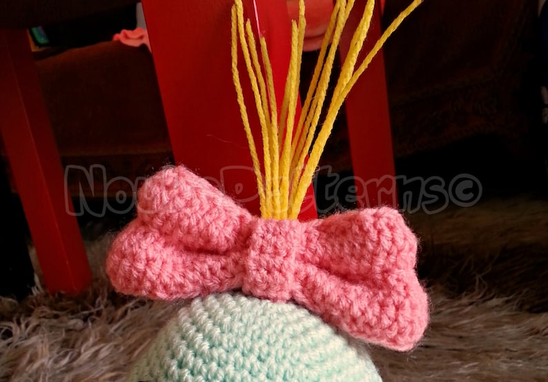 May include: A crocheted hat with a pink bow and yellow yarn strands on top. The hat is light blue and green.