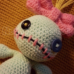May include: A crocheted blue and pink doll with a stitched mouth and button eyes. The doll has a pink bow on its head and yellow yarn hair.