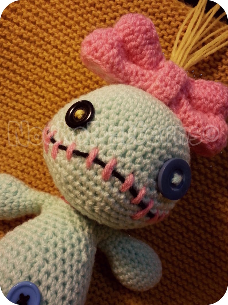 May include: A crocheted blue and pink doll with a stitched mouth and button eyes. The doll has a pink bow on its head and yellow yarn hair.