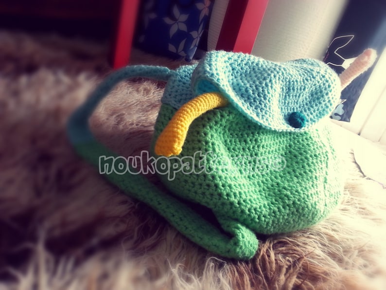 May include: A green crocheted backpack with a blue crocheted flap and a yellow crocheted strap. The backpack has a small blue pom-pom on the flap.