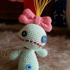 May include: A crocheted Stitch doll with a pink bow and yellow yarn hair. The doll is light blue with black stitching around the eyes and mouth. The doll has blue button eyes and a blue button on its belly.