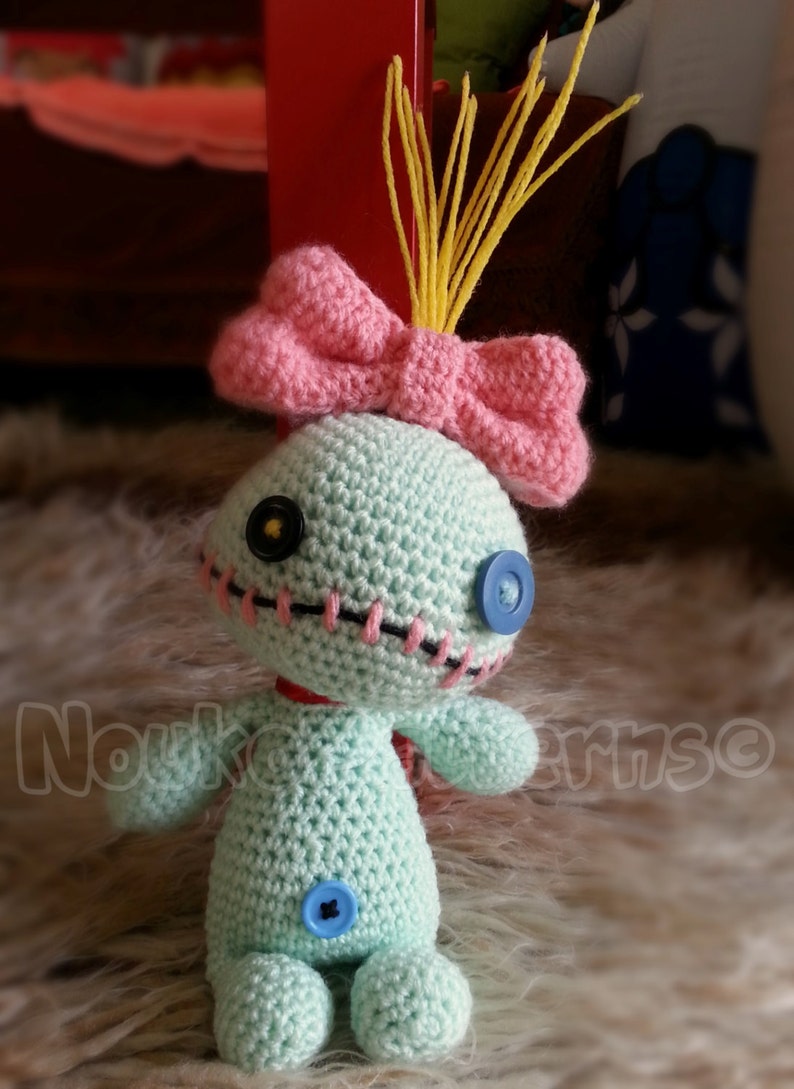 May include: A crocheted Stitch doll with a pink bow and yellow yarn hair. The doll is light blue with black stitching around the eyes and mouth. The doll has blue button eyes and a blue button on its belly.