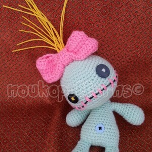 May include: A crocheted doll with a pink bow, yellow yarn hair, and black button eyes. The doll has a stitched mouth and is wearing a blue button on its belly.