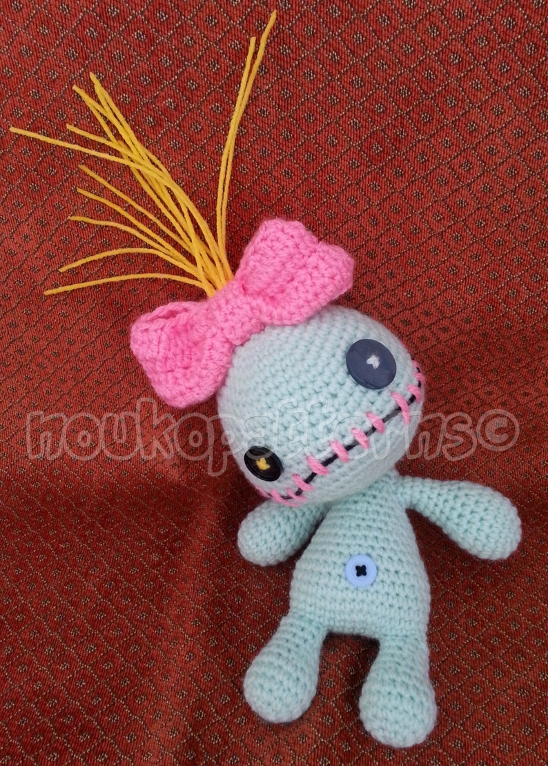 May include: A crocheted doll with a pink bow, yellow yarn hair, and black button eyes. The doll has a stitched mouth and is wearing a blue button on its belly.