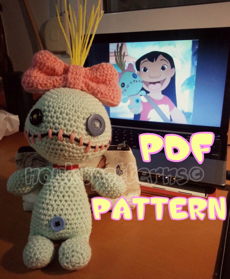 May include: A crocheted Stitch doll with a pink bow and yellow hair. The doll is sitting on a table with a laptop in the background. The laptop screen shows a scene from the movie Lilo & Stitch. The text "PDF PATTERN" is written in yellow letters on a pink background.