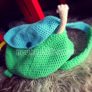 May include: A green and blue crocheted backpack with a long, pink crocheted tail. The backpack has a flap with a blue button.