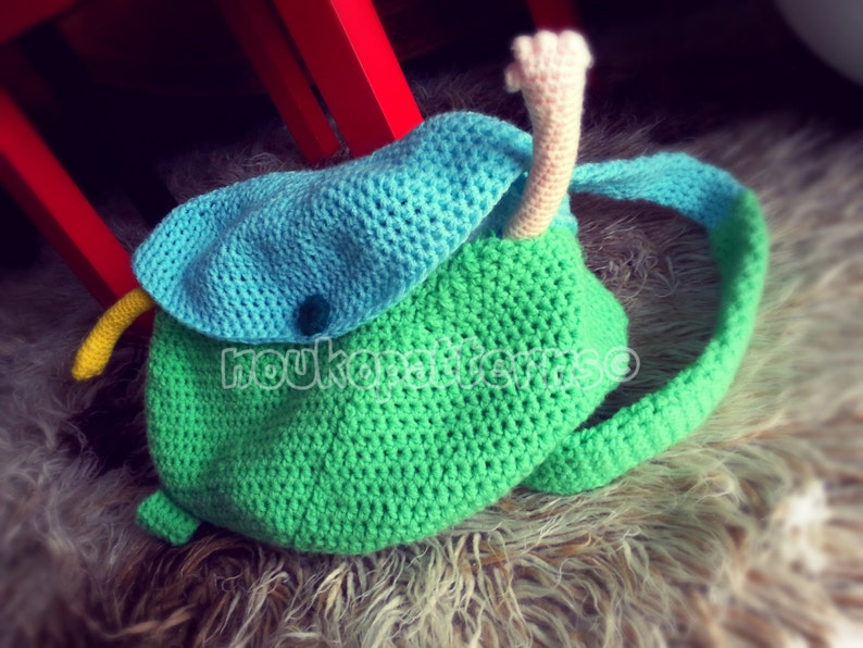 May include: A green and blue crocheted backpack with a long, pink crocheted tail. The backpack has a flap with a blue button.