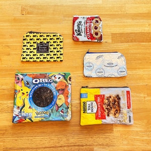 Puede incluir: Cuatro bolsas con cremallera diferentes hechas de envases de alimentos reciclados. Las bolsas son amarillas con lunares negros, azules con un diseño de Pokemon, blancas con un diseño de galletas con chispas de chocolate y blancas con un diseño de galletas de fresa. Las bolsas miden aproximadamente 10 cm de ancho y 15 cm de alto.